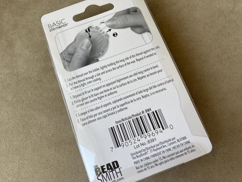 May include: A white rectangular product packaging for a thread waxer by Basic Elements and The Beadsmith. The back of the card features a black and white diagram showing how to use the tool, along with instructions in English, French, and Spanish.