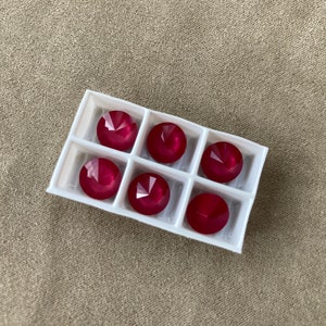 May include: Six round, faceted, ruby-red gemstones are arranged in a white, divided tray. The gems have a brilliant cut, reflecting light. The tray sits on a neutral, textured surface, creating a contrast with the vibrant stones.