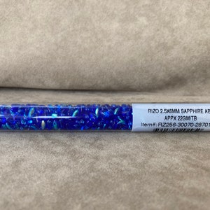 May include: A clear plastic tube filled with blue glass beads. The label reads "RIZO 2.5X6MM SAPPHIRE AB APPX 22GM/TB Item#: RZ256-30070-28701-TB"