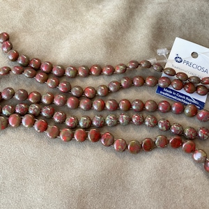 May include: A strand of red and brown speckled round beads. The beads are strung on a white thread. The beads are made in the Czech Republic. The packaging says "Preciosa Traditional Czech Beads".