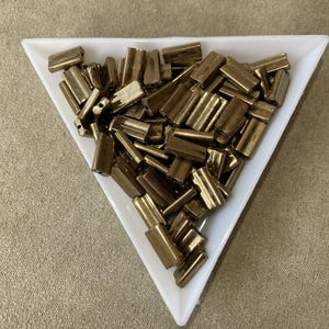May include: A collection of small, rectangular, brass-colored metal beads fills a white triangular dish. The beads have a cylindrical shape with a hole running through the center. The dish sits on a tan surface.