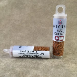 May include: Two small clear plastic tubes filled with brown seed beads. The tube on the left has a label that reads "Item#: DB2109-TB 11/0 DELICA DURACOAT OPQ SIENNA - APRX 7.2GM/TB". The tube on the right has a label that reads "MIYUKI 11/0 DELICA ΟΠ".