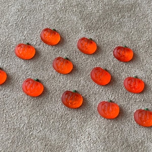 May include: A collection of orange, translucent pumpkin-shaped beads with green stems. The beads are scattered on a textured, neutral-colored surface, perfect for Halloween crafts and decorations.