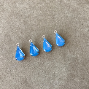 May include: Four light blue teardrop-shaped crystal pendants with silver-colored loops. The crystals have a faceted design and are set in a silver-colored metal. The pendants are displayed on a neutral-colored fabric.