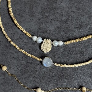 May include: Three necklaces with different designs. The top necklace has small wooden beads. The middle necklace features a carved hand pendant, pearls, and silver beads. The bottom necklace has a moonstone pendant and a gold chain.