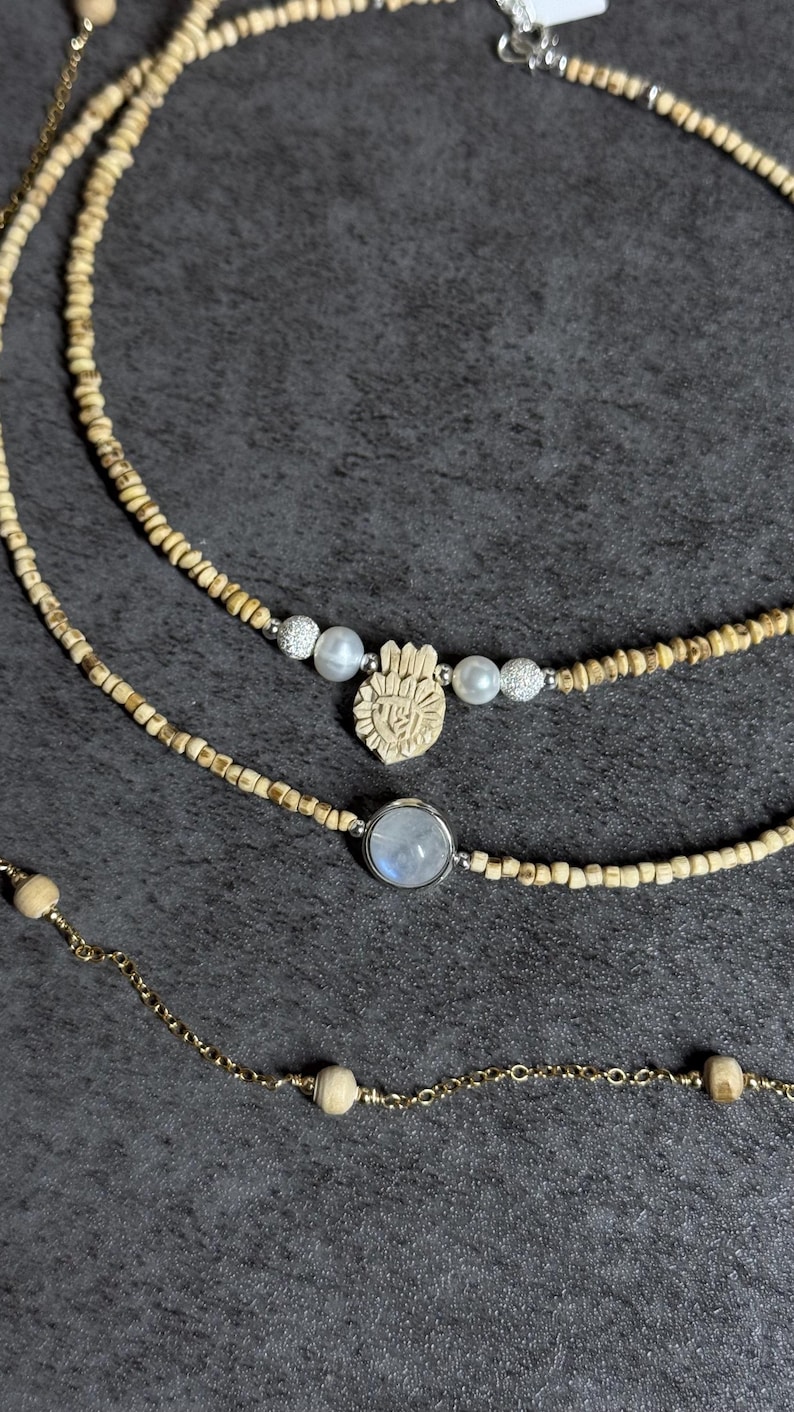May include: Three necklaces with different designs. The top necklace has small wooden beads. The middle necklace features a carved hand pendant, pearls, and silver beads. The bottom necklace has a moonstone pendant and a gold chain.