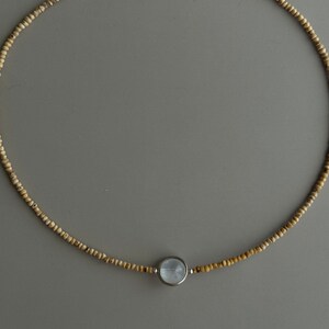 May include: A delicate necklace featuring small, light brown beads strung together. The necklace has a silver clasp and a central pendant with a round, light blue stone set in a silver bezel. The necklace is displayed on a gray background.