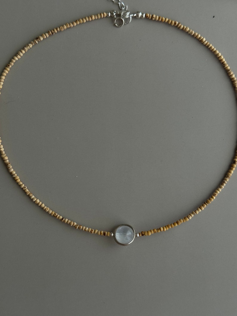 May include: A delicate necklace featuring small, light brown beads strung together. The necklace has a silver clasp and a central pendant with a round, light blue stone set in a silver bezel. The necklace is displayed on a gray background.