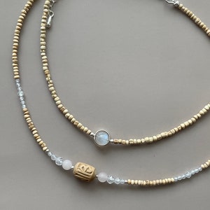 May include: Two layered necklaces. The top necklace features small, light-colored beads and a silver pendant with a round, light-colored stone. The bottom necklace has similar beads, clear crystal accents, and a carved wooden bead.