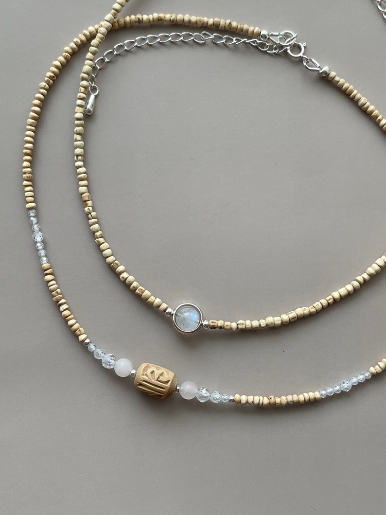 May include: Two layered necklaces. The top necklace features small, light-colored beads and a silver pendant with a round, light-colored stone. The bottom necklace has similar beads, clear crystal accents, and a carved wooden bead.