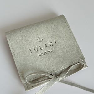 May include: A light gray suede pouch with a matching tie closure. The pouch features the embossed text "TULASI AND PEARLS" in a simple, elegant font. The pouch is designed to hold jewelry or small items.