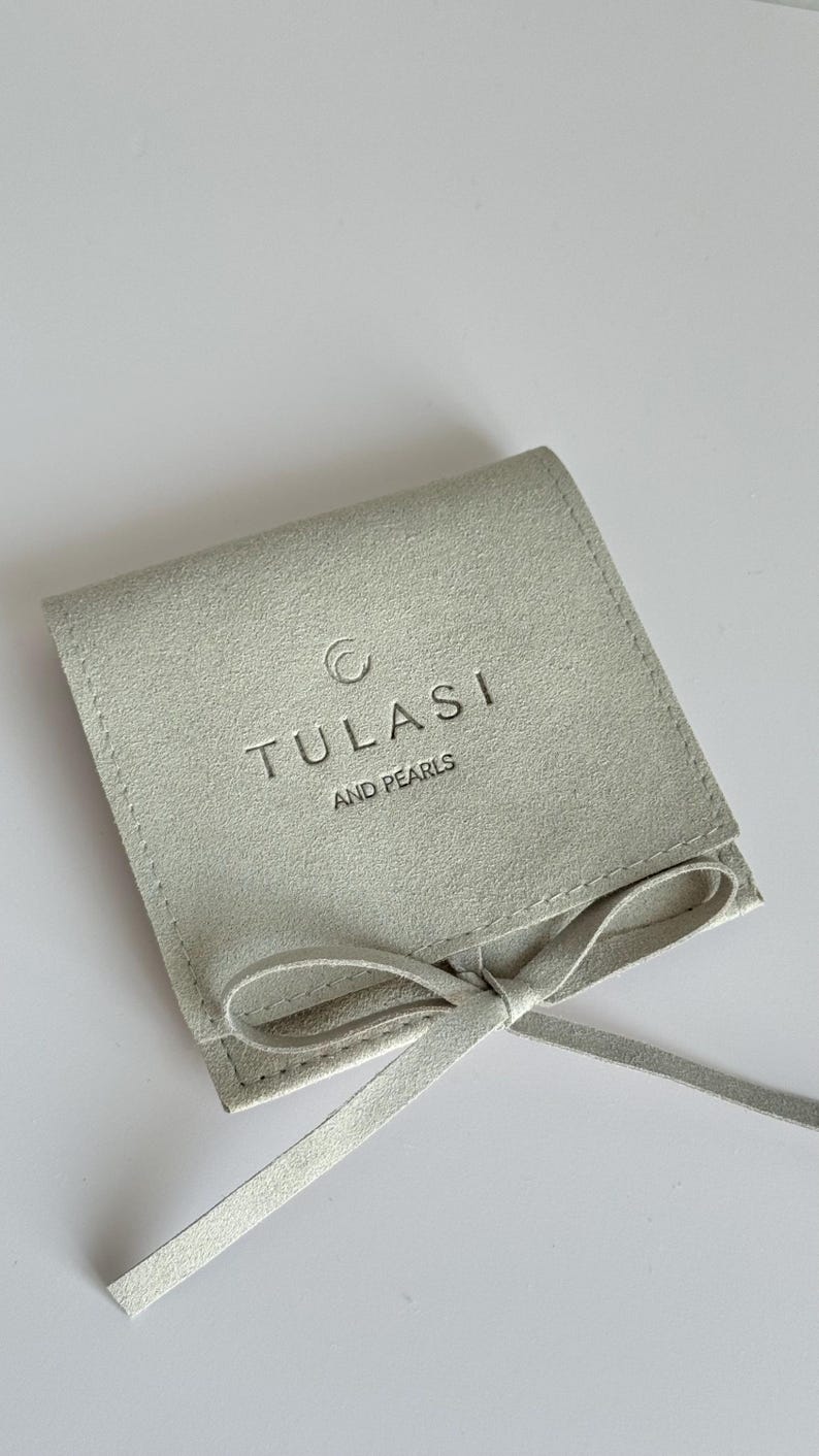May include: A light gray suede pouch with a matching tie closure. The pouch features the embossed text "TULASI AND PEARLS" in a simple, elegant font. The pouch is designed to hold jewelry or small items.