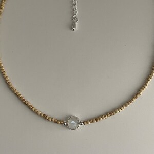 May include: A delicate necklace featuring small, light brown wooden beads. A silver-toned pendant holds a round, iridescent stone. The necklace has a silver-toned adjustable chain. This piece is suitable for everyday wear.