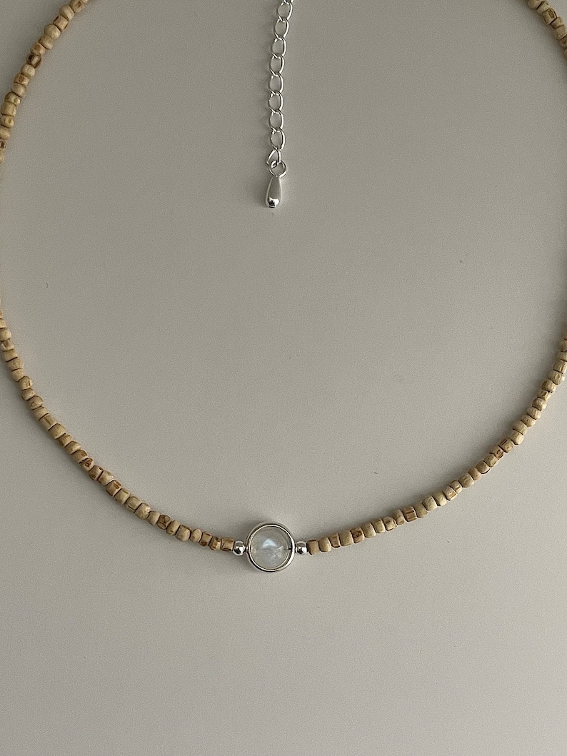 May include: A delicate necklace featuring small, light brown wooden beads. A silver-toned pendant holds a round, iridescent stone. The necklace has a silver-toned adjustable chain. This piece is suitable for everyday wear.