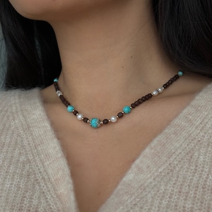 Puede incluir: Un collar de cuentas marrones, turquesas y blancas con un acento dorado. El collar está hecho de cuentas pequeñas y redondas y tiene un aspecto delicado y femenino.