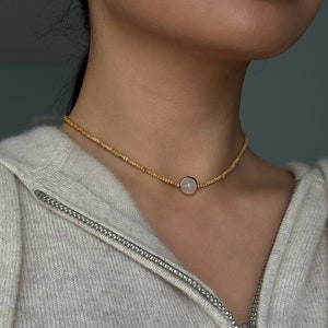 May include: A delicate necklace featuring a small, round, pale pink gemstone pendant set in a gold-colored metal. The necklace is composed of small, light brown beads. The person is wearing a light gray zip-up hoodie.