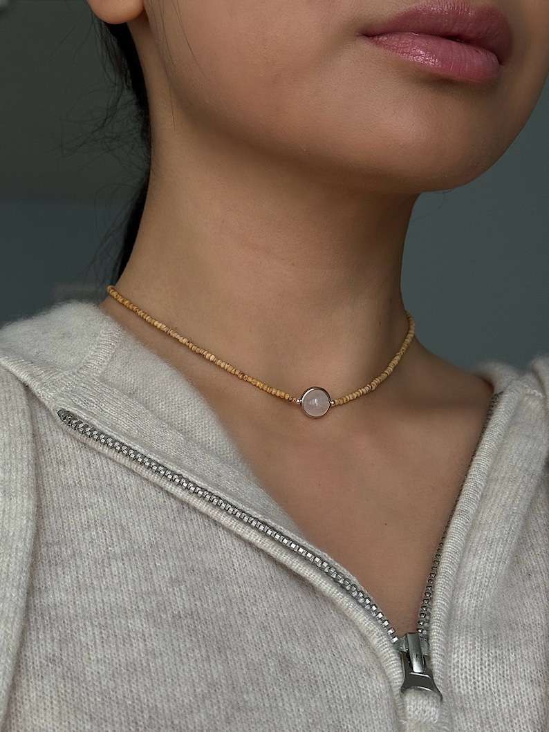 May include: A delicate necklace featuring a small, round, pale pink gemstone pendant set in a gold-colored metal. The necklace is composed of small, light brown beads. The person is wearing a light gray zip-up hoodie.