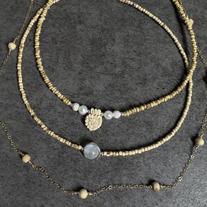 May include: Three layered necklaces with different designs. The top necklace has small, light-colored beads. The middle necklace features a hand-shaped pendant with pearls and crystals. The bottom necklace has a round, light-colored stone pendant.
