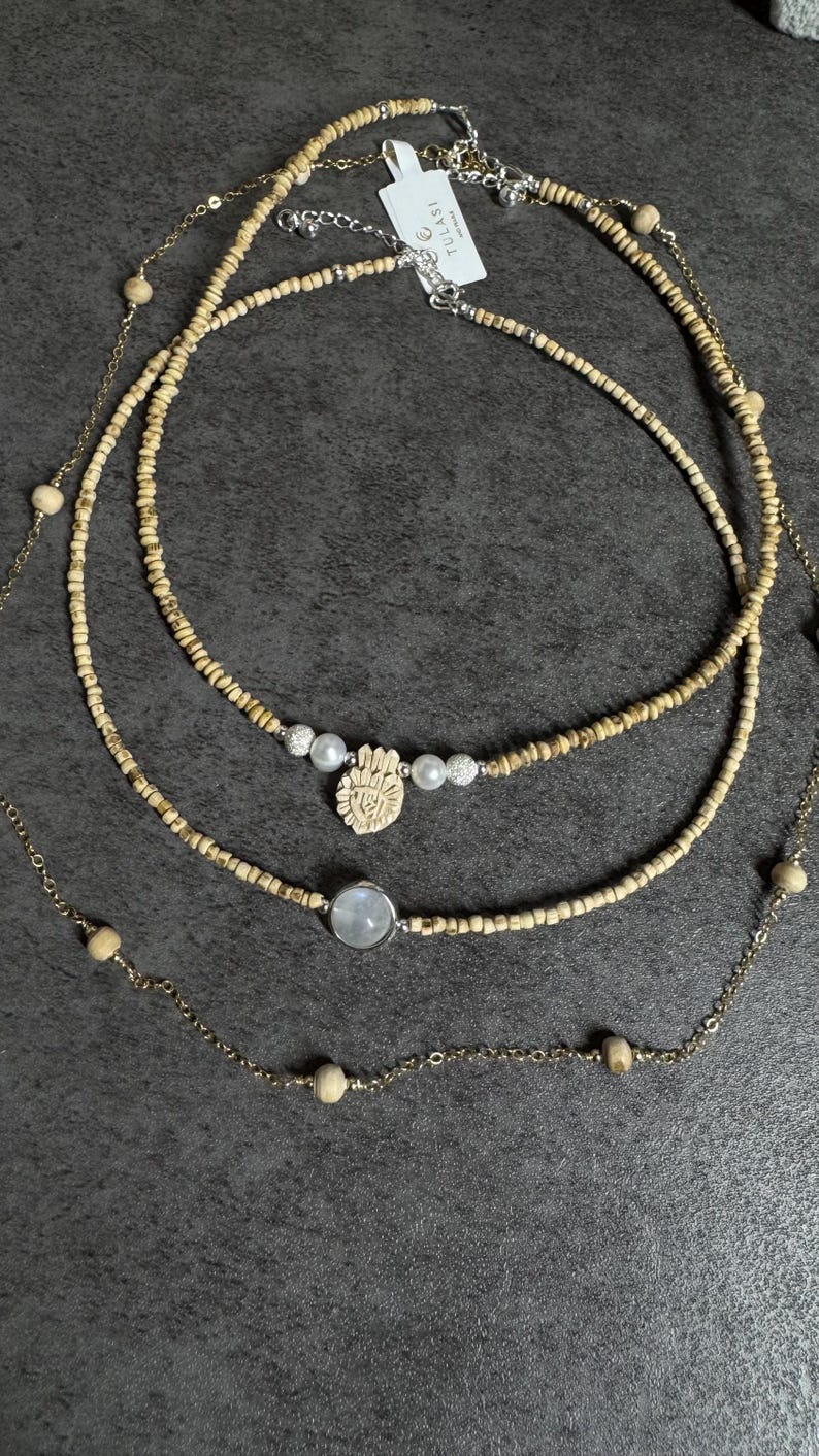 May include: Three layered necklaces with different designs. The top necklace has small, light-colored beads. The middle necklace features a hand-shaped pendant with pearls and crystals. The bottom necklace has a round, light-colored stone pendant.