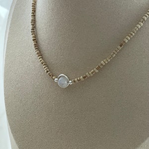 May include: A delicate necklace featuring small, light brown beads strung together. A silver-toned pendant with a round, pale blue stone is centered on the necklace. The necklace is displayed on a neutral-toned mannequin.