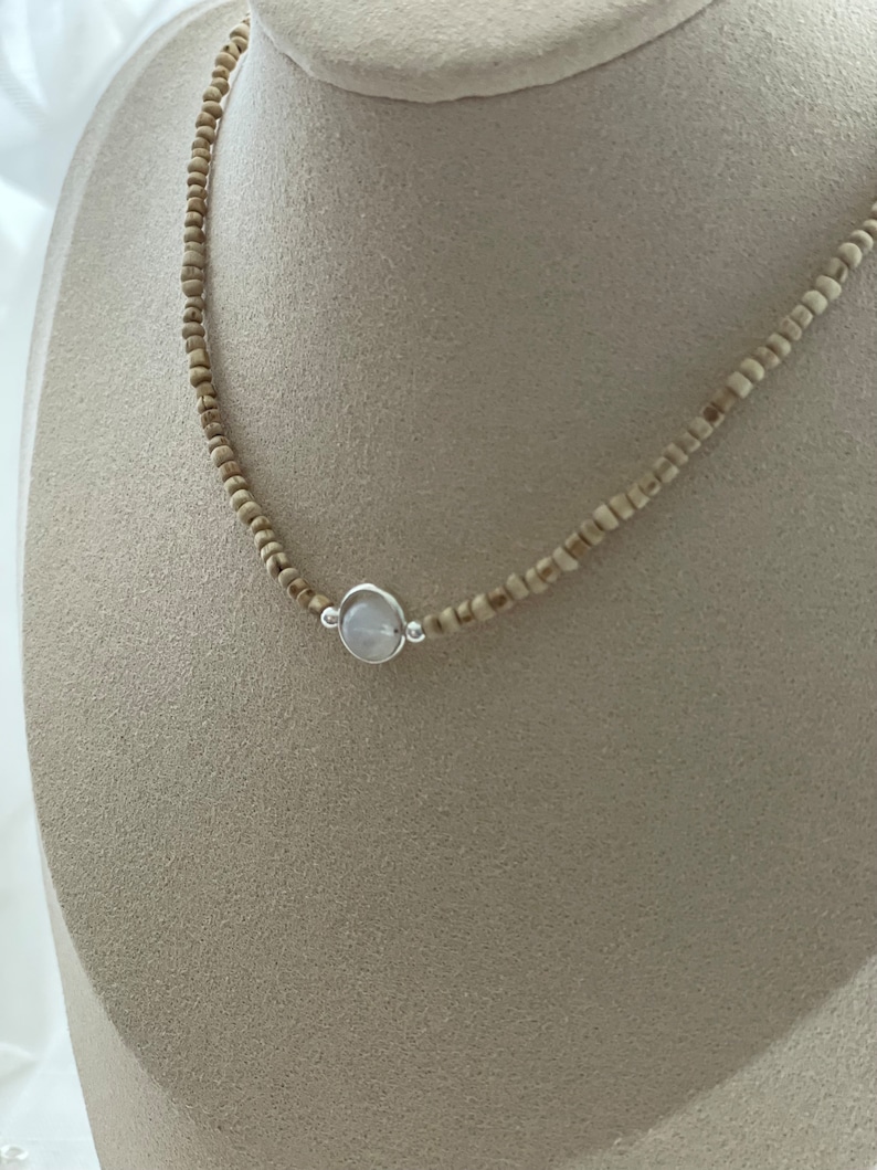 May include: A delicate necklace featuring small, light brown beads strung together. A silver-toned pendant with a round, pale blue stone is centered on the necklace. The necklace is displayed on a neutral-toned mannequin.