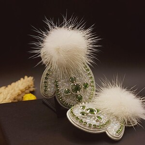 Pode incluir: Dois broches de pompom de pele branca com designs de contas verdes e brancas. Os broches têm a forma de luvas.