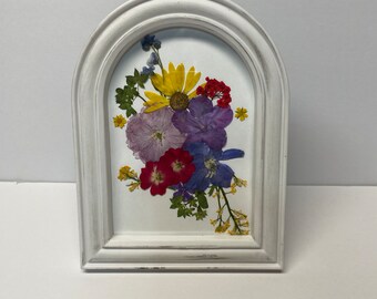 Könnte beinhalten: Ein weißer, gewölbter Rahmen mit einem gepressten Blumenarrangement darin. Die Blumen sind in den Farben Lila, Rot, Gelb und Blau.