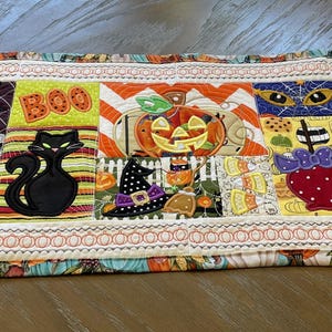 May include: A rectangular quilted table runner featuring a patchwork design with Halloween motifs including white ghosts, a black cat, a jack-o'-lantern, the word BOO, a witch hat, a candy apple, a crescent moon, a bat, and striped witch boots.