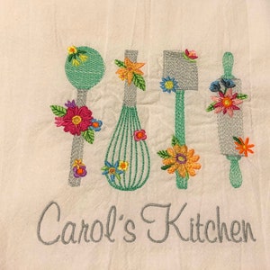May include: Embroidered kitchen towel with a green whisk, a gray spatula, a gray rolling pin, and a gray spoon. Each utensil is decorated with colorful flowers. The text "Carol's Kitchen" is embroidered in gray at the bottom of the towel.