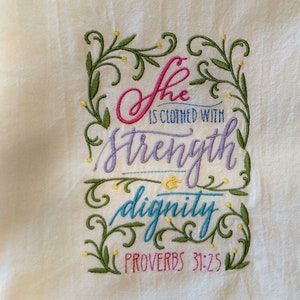 May include: Cream-colored embroidered towel with the text "She is clothed with strength dignity PROVERBS 31:25." The text is in various colors, including pink, purple, and teal, and is surrounded by green and yellow floral embroidery.