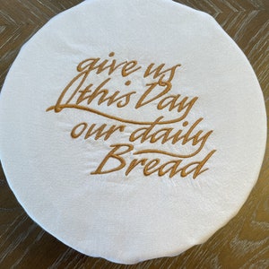 May include: White round cloth bread cover with gold embroidery that reads "give us this day our daily bread".