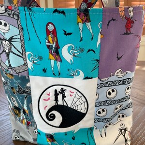 May include: A blue and white patchwork tote bag with a Nightmare Before Christmas theme. The bag features characters like Jack Skellington, Sally, and Oogie Boogie. The bag has a large embroidered circle with Jack Skellington and Sally standing on his head.
