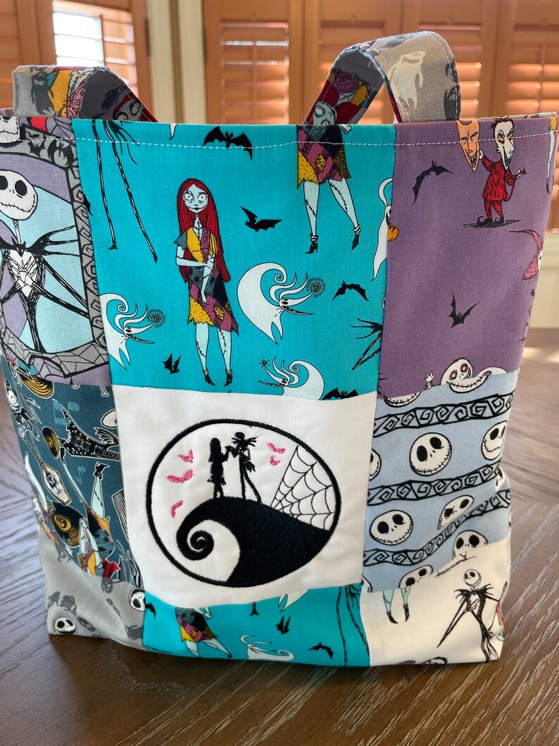 May include: A blue and white patchwork tote bag with a Nightmare Before Christmas theme. The bag features characters like Jack Skellington, Sally, and Oogie Boogie. The bag has a large embroidered circle with Jack Skellington and Sally standing on his head.