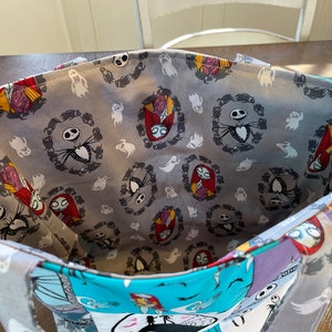 May include: A gray tote bag with a Nightmare Before Christmas themed fabric lining. The lining features a repeating pattern of Jack Skellington and Sally in wreaths, surrounded by ghosts.