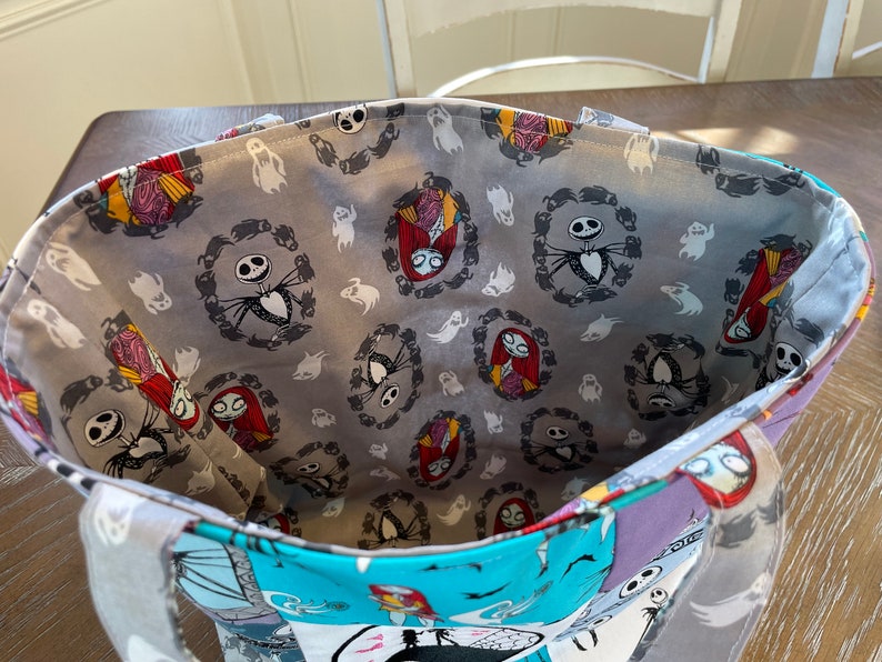 May include: A gray tote bag with a Nightmare Before Christmas themed fabric lining. The lining features a repeating pattern of Jack Skellington and Sally in wreaths, surrounded by ghosts.