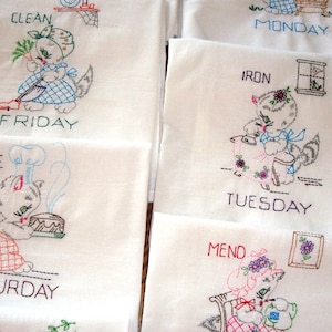 May include: Embroidered white fabric squares with cartoon cat illustrations depicting daily chores. Each square features a cat with text indicating the day of the week: Friday, Monday, Tuesday, Saturday, and Mending. The cats are in various colors.