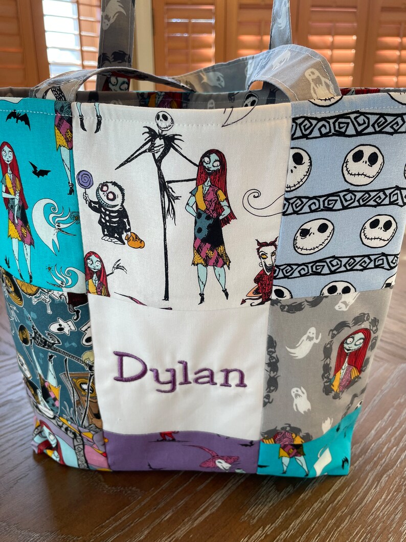 May include: A patchwork tote bag with a Nightmare Before Christmas theme. The bag is made of different fabrics with various characters from the movie, including Jack Skellington and Sally. The bag has a white panel with the name "Dylan" embroidered in purple.
