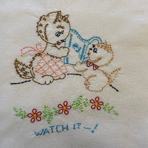 May include: Embroidered kitchen towel with two kittens, one holding a blue pitcher and the other with a pink bow. The towel has a green vine with red flowers and the text "WATCH IT!" embroidered on it.