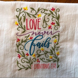 May include: White kitchen towel with embroidered floral border and the text "LOVE never fails 1 CORINTHIANS 13:8" in colorful lettering.