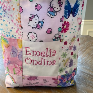 May include: A pink and white patchwork tote bag with Hello Kitty and butterfly designs. The bag has a white panel with the name "Emeli Ondina" embroidered in pink.
