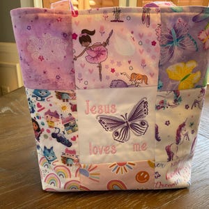 May include: A patchwork tote bag with various patterns. The bag features panels with ballerinas, butterflies, and unicorns. A central panel displays a butterfly and the text "Jesus loves me". The bag has a pink and purple color scheme.