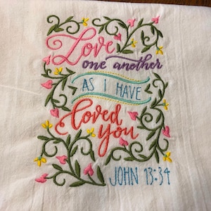 May include: White kitchen towel with embroidered floral design and the text "Love one another as I have loved you John 13:34".