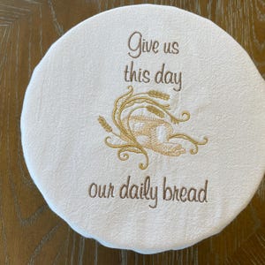 May include: A round, off-white bread warmer with embroidered text and wheat design. The text reads "Give us this day our daily bread" in brown thread. The wheat design is in gold thread.