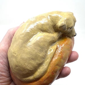 May include: A ceramic sculpture of a dog curled up in a loaf of bread. The dog is light brown and the bread is orange.