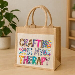 May include: A beige jute tote bag with a white front panel. The panel displays the text "CRAFTING IS MY THERAPY" in colourful, patterned letters, alongside illustrations of crafting tools such as scissors and yarn. The bag has two strong handles.