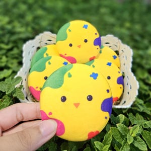 Pode incluir: Uma coleção de itens amarelos, redondos, de pelúcia em forma de pintinho com detalhes coloridos em verde, roxo, rosa e azul. Os itens estão em uma cesta branca, com um sendo segurado por uma mão. O fundo é uma folhagem verde desfocada.