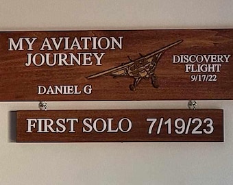 May include: Two wooden signs with white text. The top sign reads "My Aviation Journey, Daniel G, Discovery Flight 9/17/22". The bottom sign reads "First Solo 7/19/23".