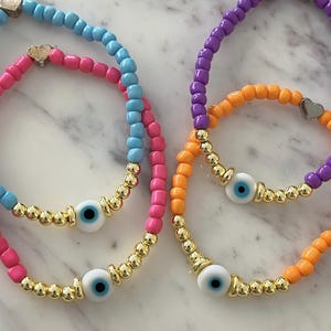 May include: Three colorful beaded bracelets with gold accents and evil eye charms. The bracelets are pink and blue, orange and purple, and orange and purple.