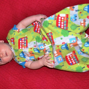 May include: A baby wearing a green and red sleep sack with a bus pattern. The sleep sack has a white zipper and is made of soft fabric.