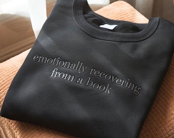 Puede incluir: Una sudadera negra de cuello redondo con el texto bordado "emotionally recovering from a book". La sudadera está doblada y apoyada sobre un otomán de pana. La imagen está iluminada por luz natural.